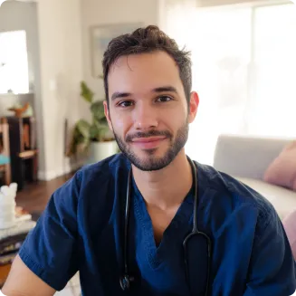 A young male healthcare professional in navy scrubs with a stethoscope around his neck smiles warmly while seated in a cozy, sunlit room.