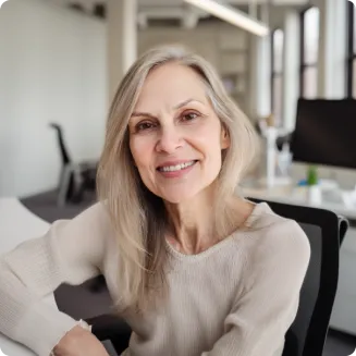 A smiling woman with long, light gray hair sits in a modern office, looking warmly at the camera.