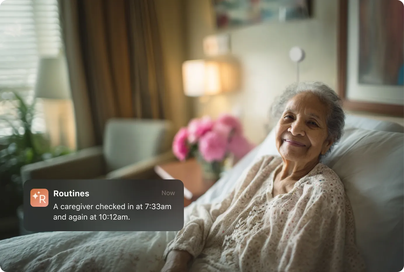 A smiling elderly woman relaxes in bed with a white sensor on the all behind her, as a Routines app notification confirms her caregiver checked in at 7:33 a.m. and again at 10:12 a.m.